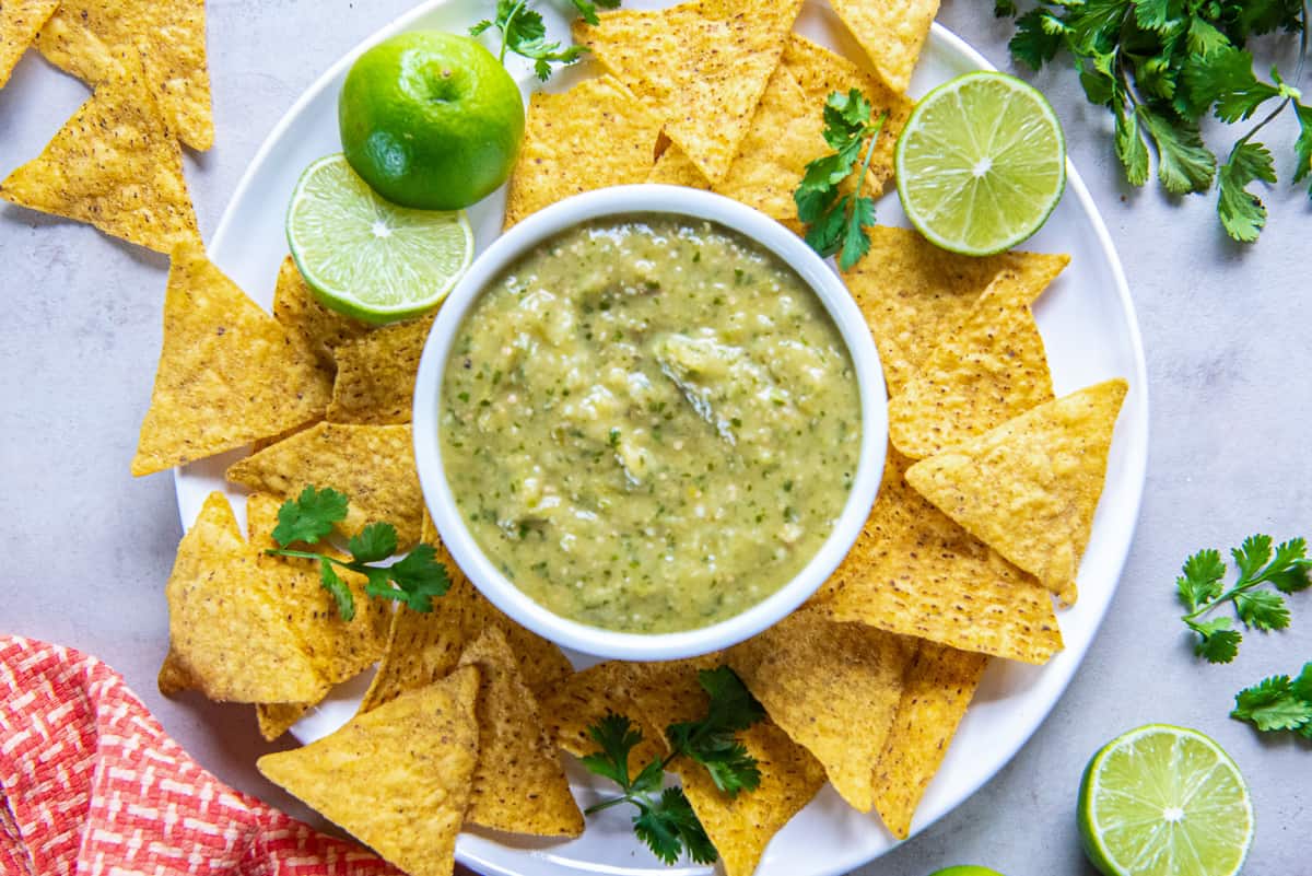 A top down shot of a small bowl of tomatillo salsa on a plate with tortilla chips, lime halves, and sprigs of cilantro.