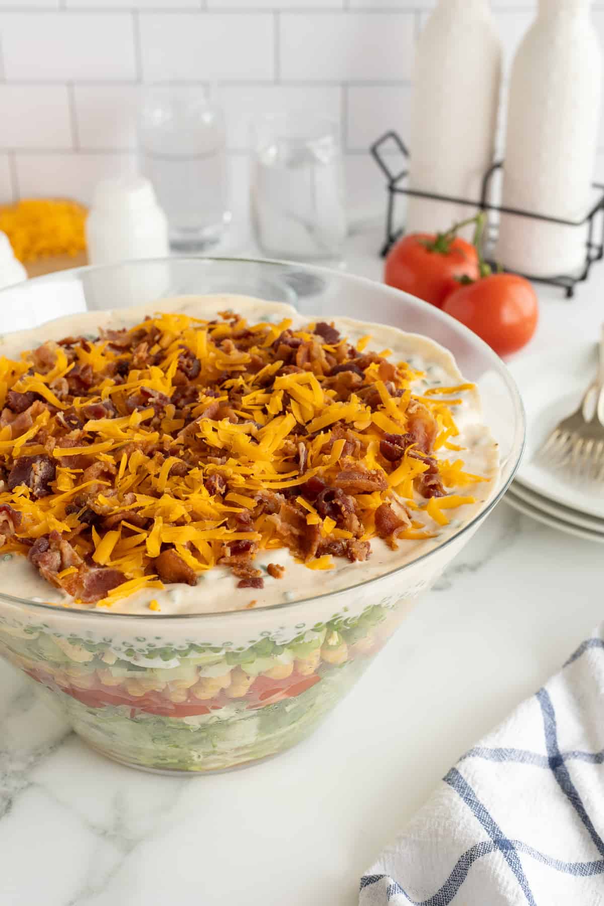 A layered salad in a glass dish topped with shredded cheddar cheese and crumbled bacon on a kitchen counter.