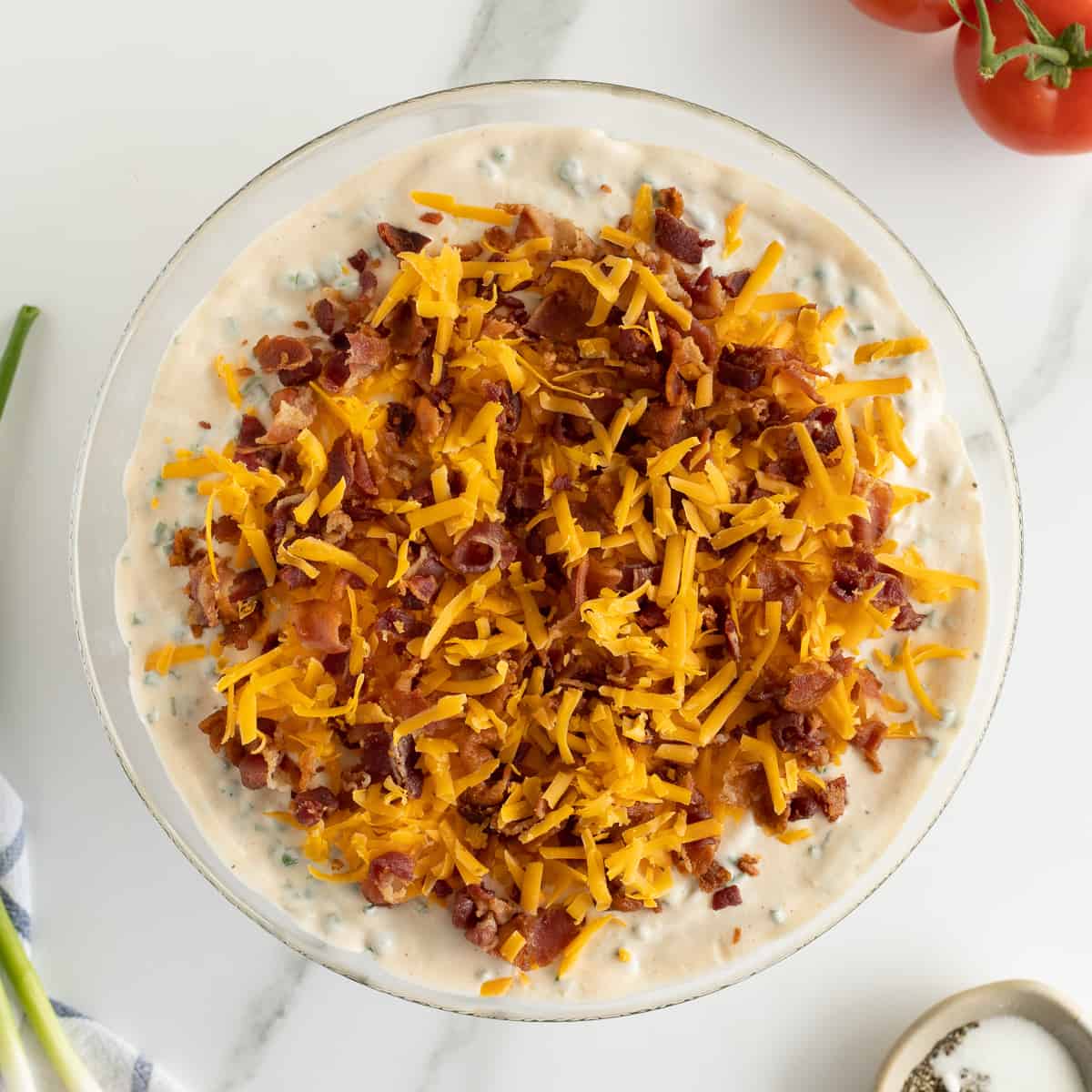 A garnish of shredded cheddar cheese and crumbled bacon on top of a seven layer salad in a glass bowl.