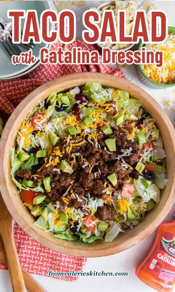 A top down shot of taco salad in a wood salad bowl next to a bottle of Catalina dressing, a small bowl of cheese, and crushed tortilla chips with text.