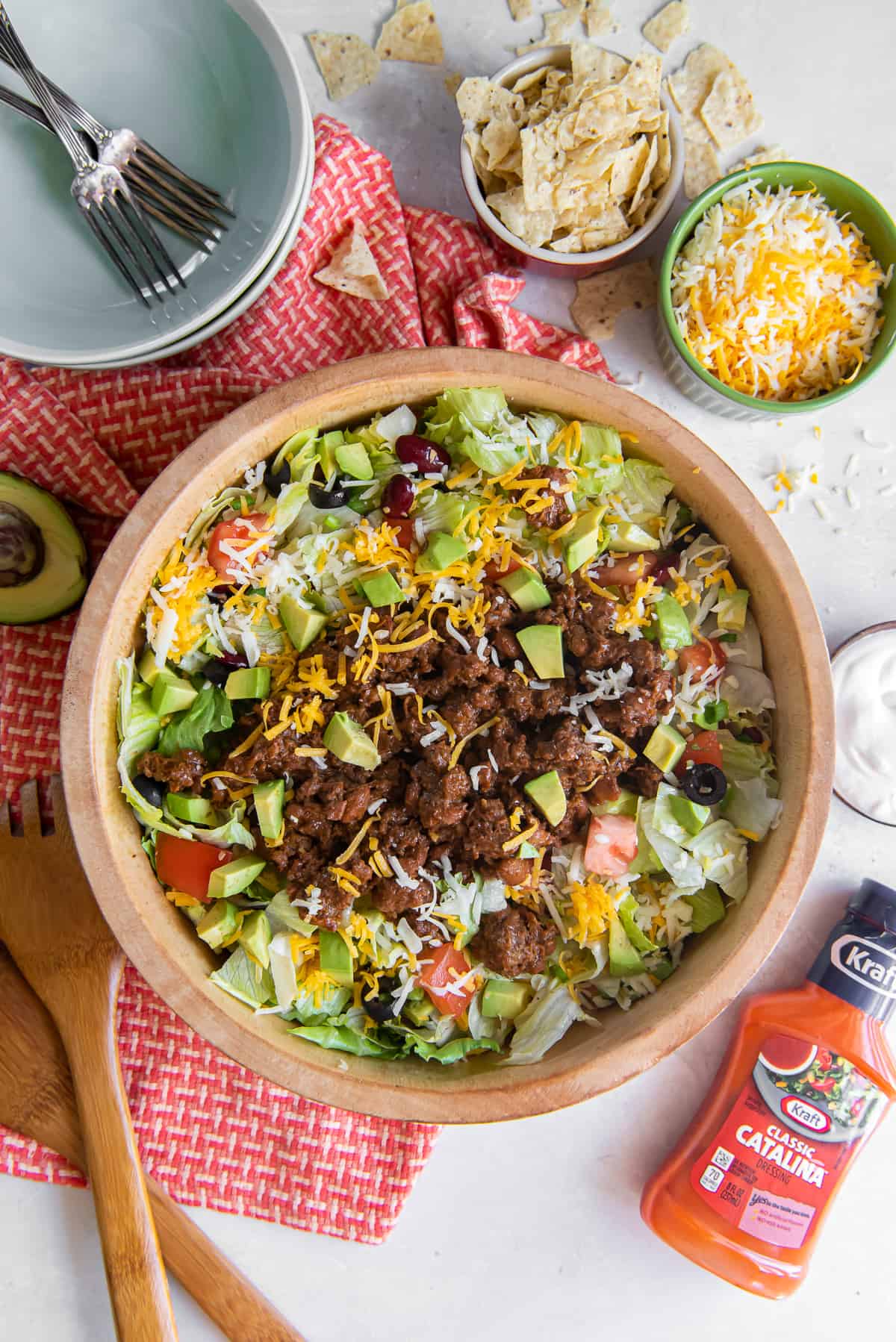 A top down shot of taco salad in a wood salad bowl next to a bottle of Catalina dressing, a small bowl of cheese, and crushed tortilla chips.