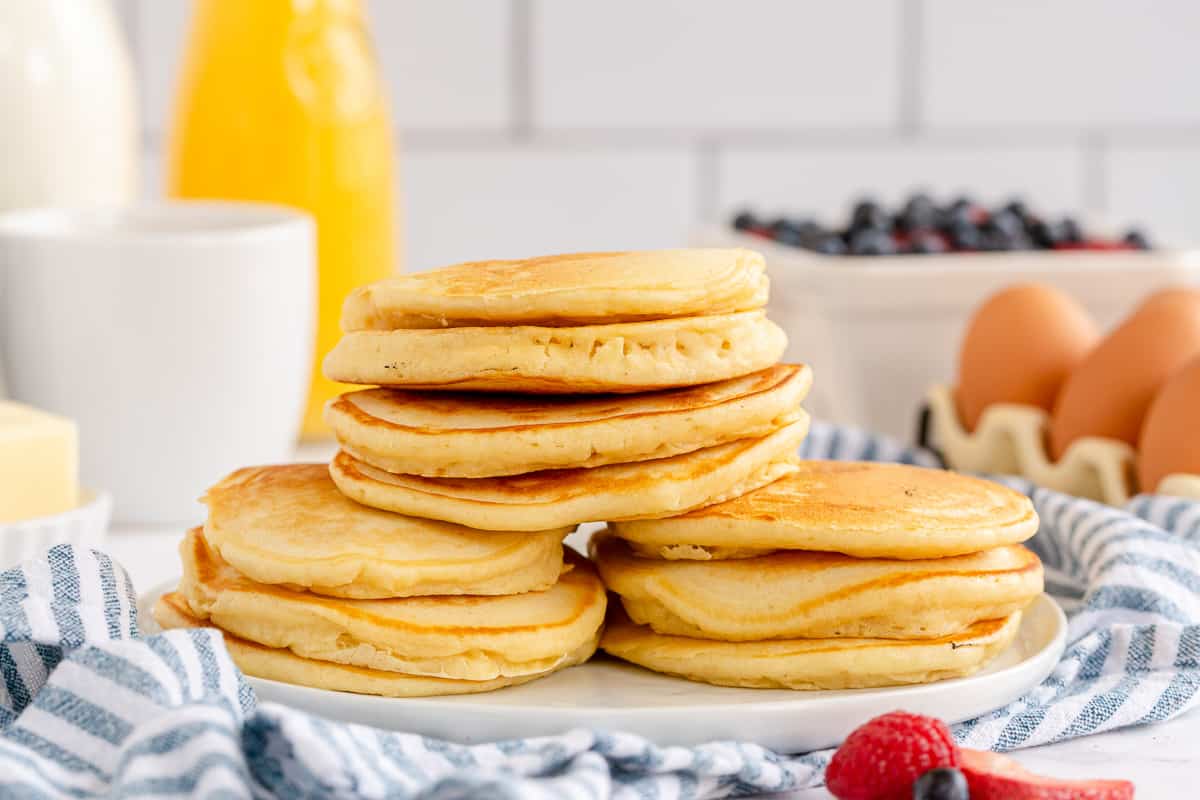 Three stacks of pancakes on a white platter in front of a glass pitcher of orange juice, carton of eggs, and fresh berries.