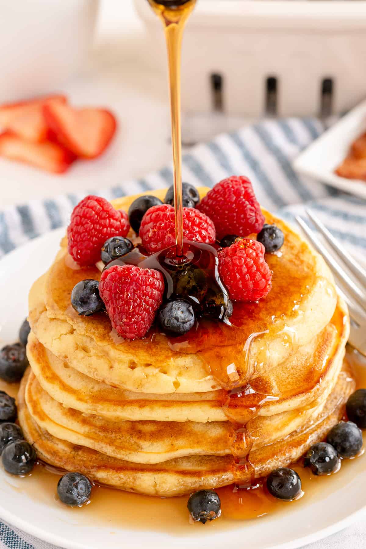 Maple syrup pouring over a stack of old fashioned pancakes topped with berries.