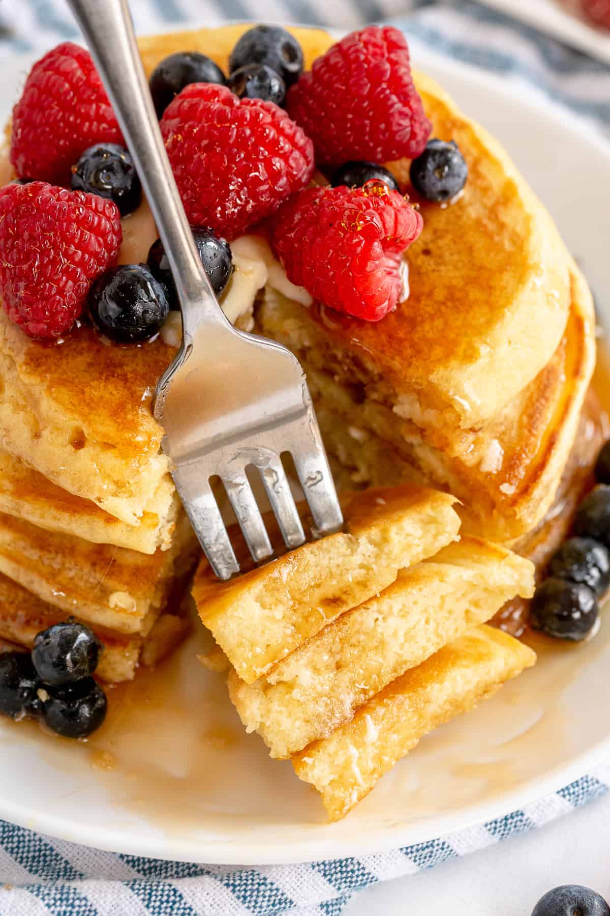 A fork piercing pieces of pancake on a plate of a stack of pancakes.