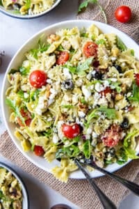 A top down shot of serving spoons resting in a large serving bowl filled with chicken pesto pasta salad.