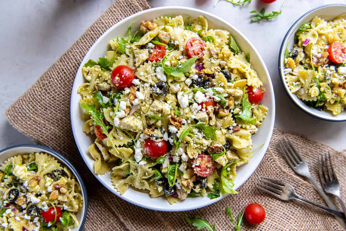 A top down shot of a large serving bowl filled with chicken pesto pasta salad next to two small serving bowls.