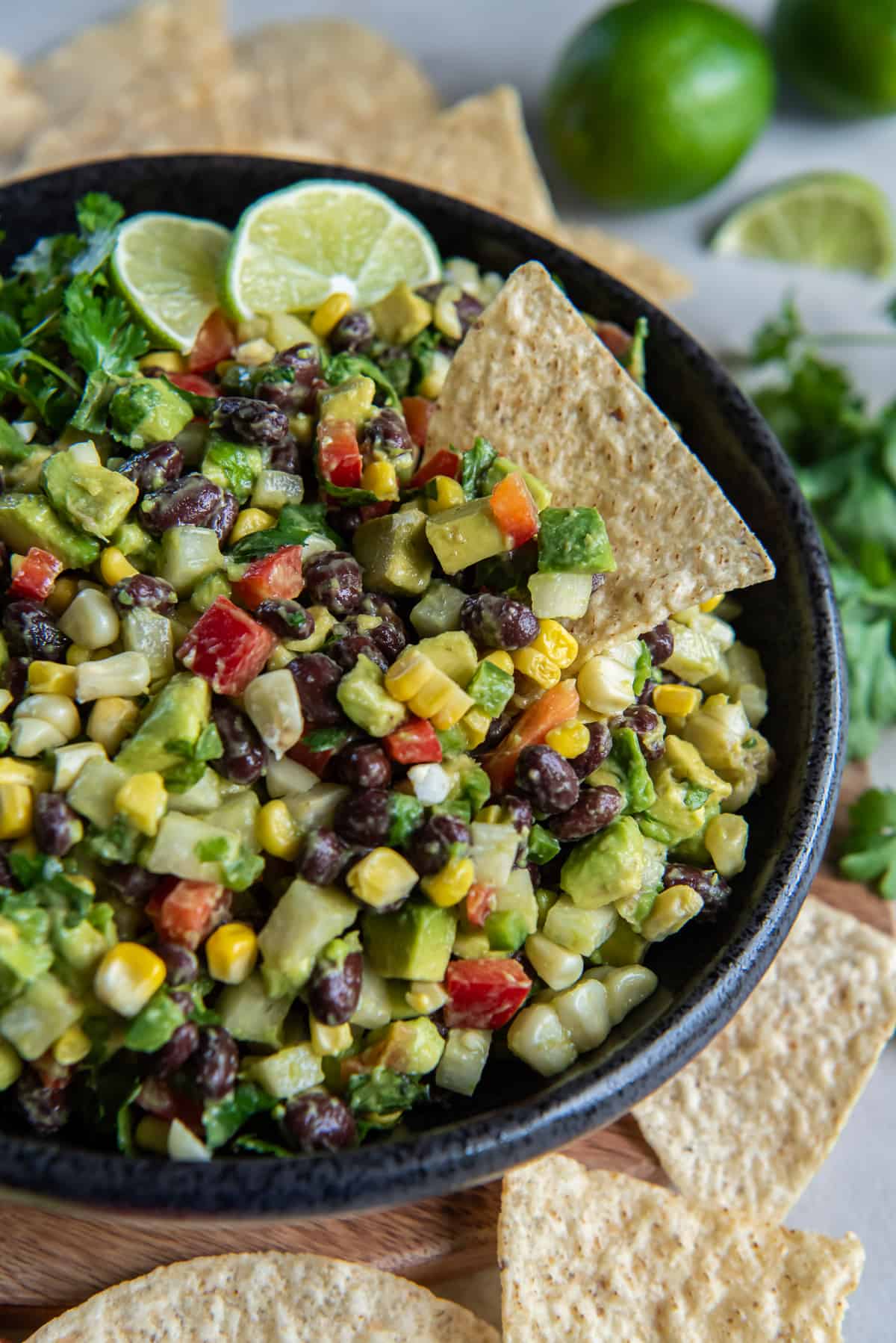 Guacamole with black bans, corn, jicama, and other ingredients in a black bowl with lime wedges and a tortilla chip.