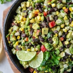 A top down shot of a guacamole loaded with black beans, red pepper, corn, and other ingredients in a black bowl with tortilla chips and cilantro sprigs around it.