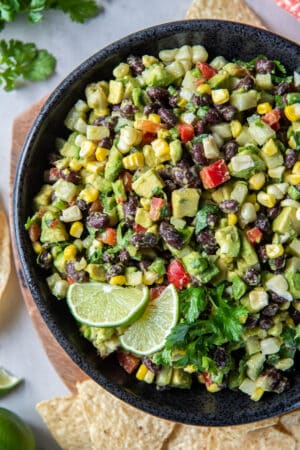 A top down shot of a guacamole loaded with black beans, red pepper, corn, and other ingredients in a black bowl with tortilla chips and cilantro sprigs around it.