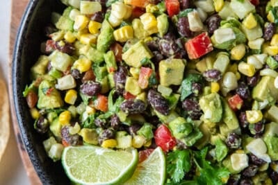 A top down shot of a guacamole loaded with black beans, red pepper, corn, and other ingredients in a black bowl with tortilla chips and cilantro sprigs around it.