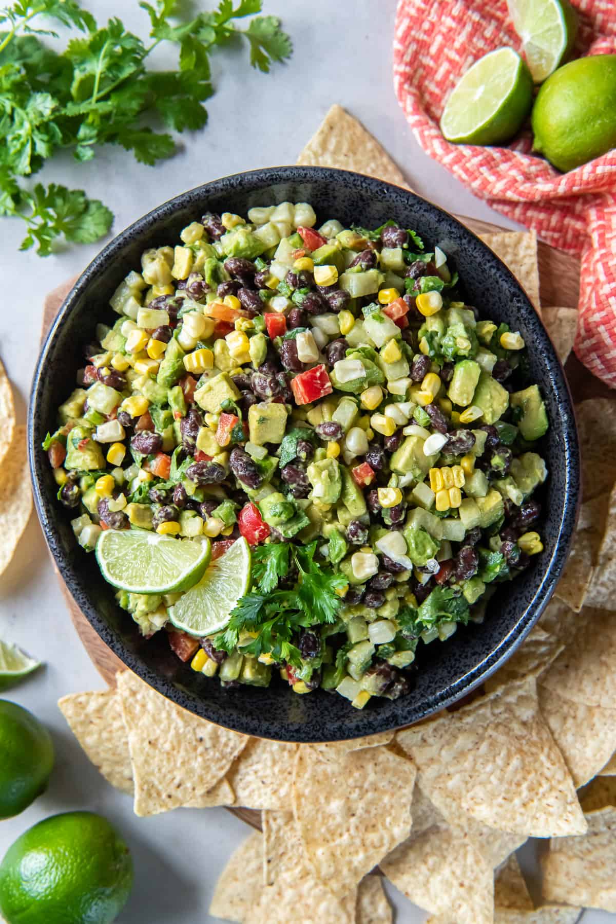 A top down shot of a guacamole with black beans, red pepper, corn and other ingredients in a black bowl with tortilla chips, cilantro sprigs and lime wedges around it.