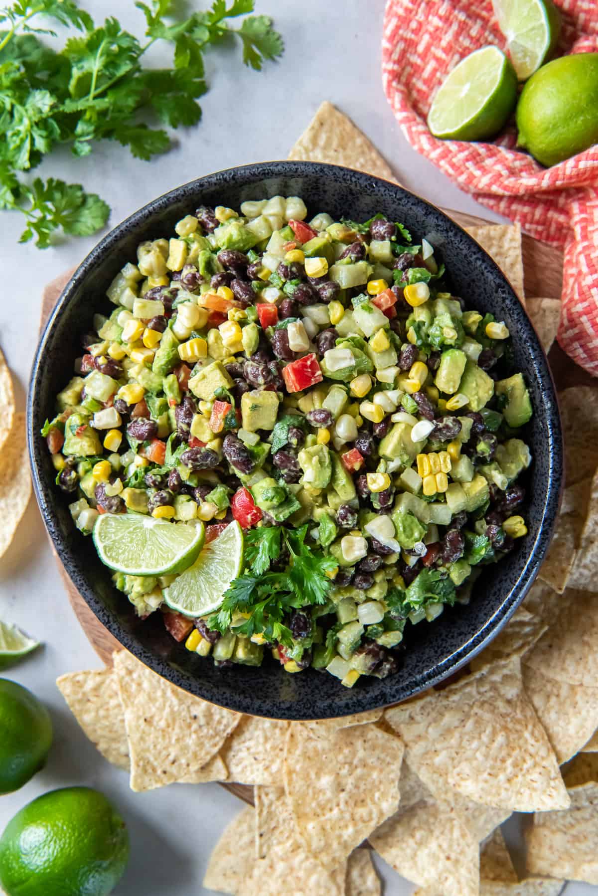 A top down shot of a guacamole loaded with black beans, red pepper, corn, and other ingredients in a black bowl with tortilla chips, cilantro sprigs, and lime wedges around it.