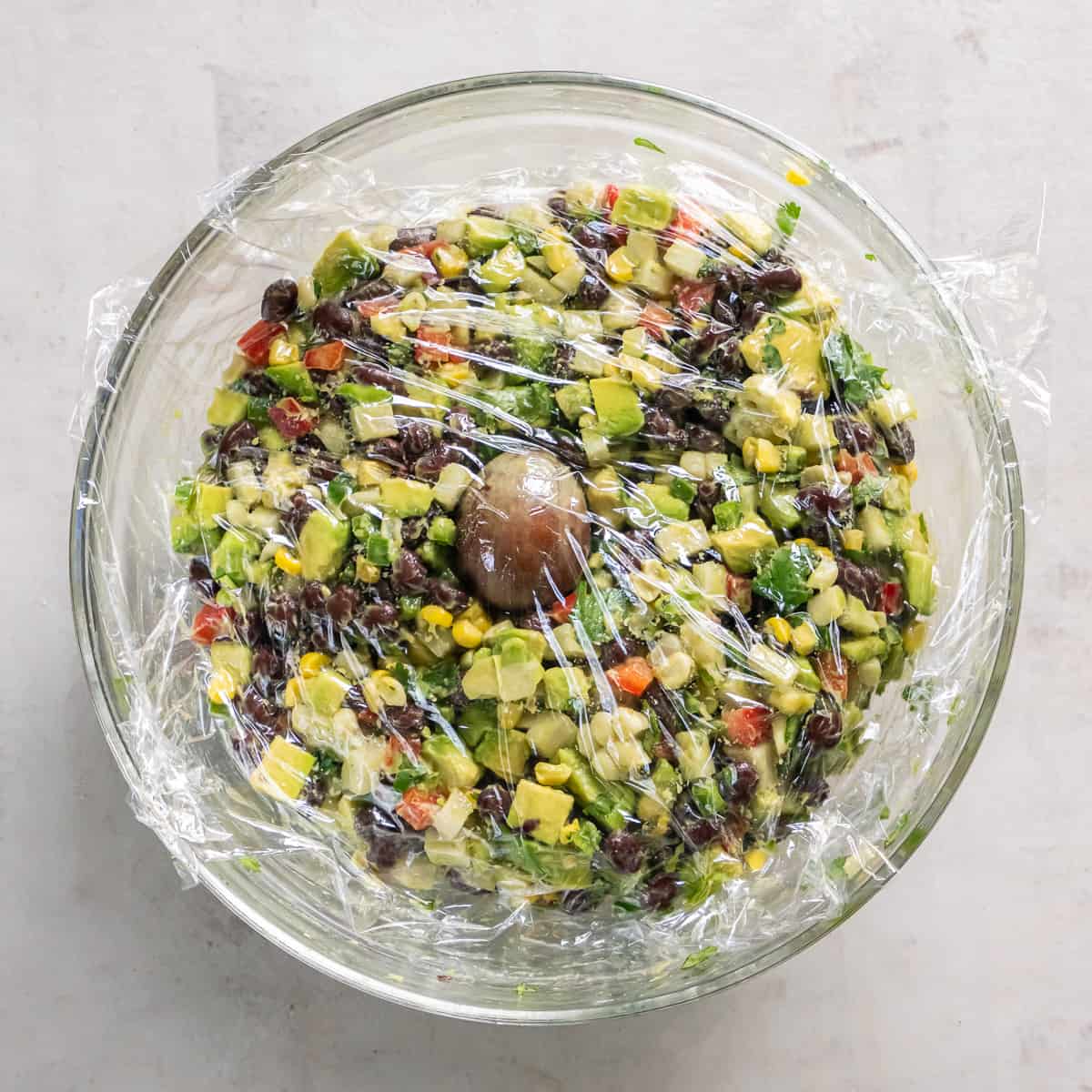 A bowl of guacamole with an avocado pit pressed into it and covered tightly with plastic wrap.