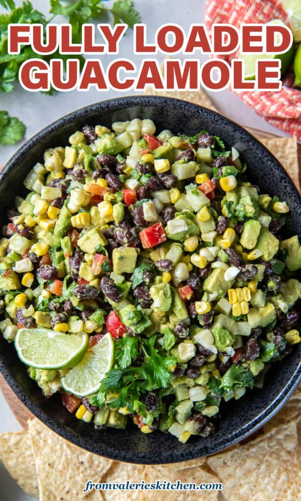 A top down shot of a guacamole loaded with black beans, red pepper, corn, and other ingredients in a black bowl with tortilla chips, cilantro sprigs, and lime wedges around it with text.