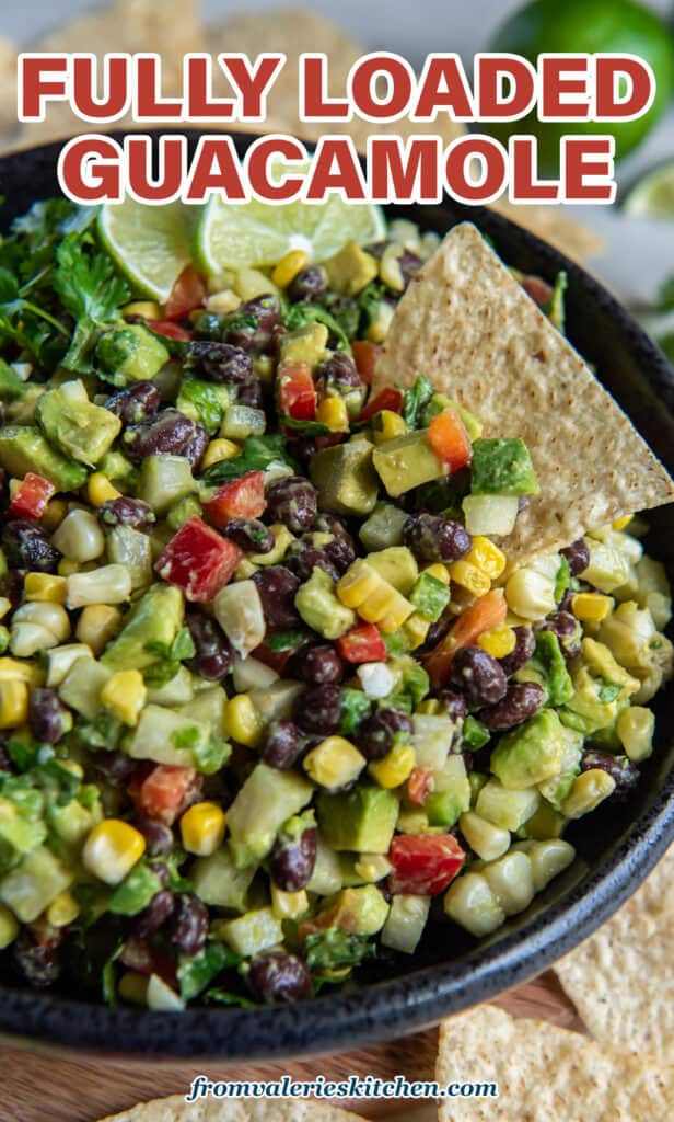Guacamole with black bans, corn, jicama, and other ingredients in a black bowl with lime wedges and a tortilla chip with text.