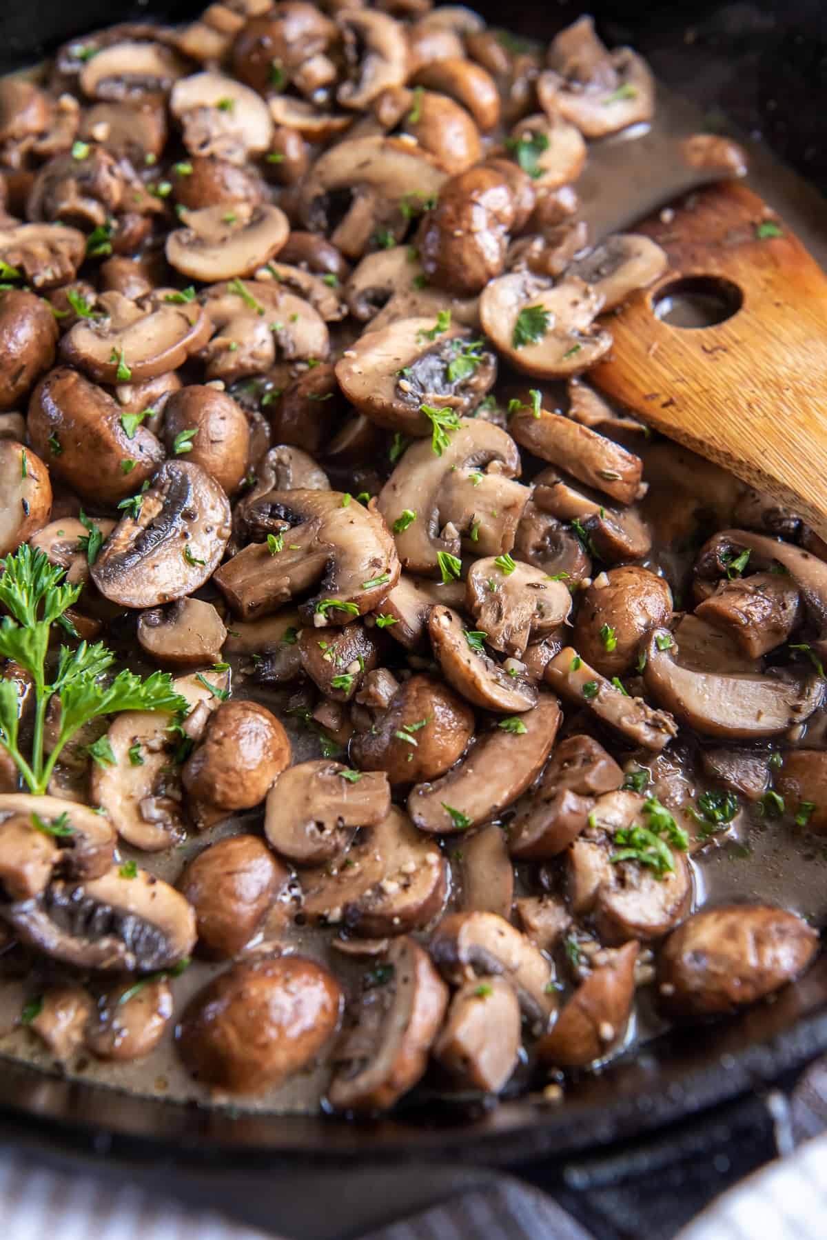 A closeup of sautéed mushrooms in a cast iron skillet with a wooden spoon.