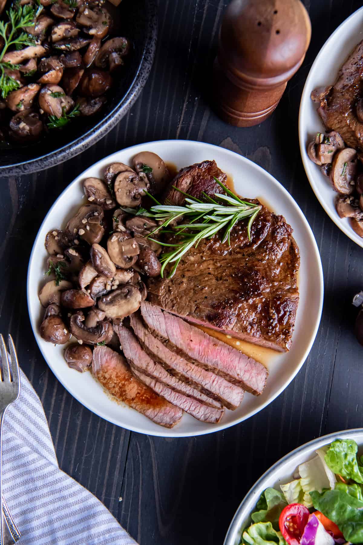 A top down shot of a dinner plate with sliced steak and mushrooms surrounded by a bowl of salad and a bowl of sauteed mushrooms.