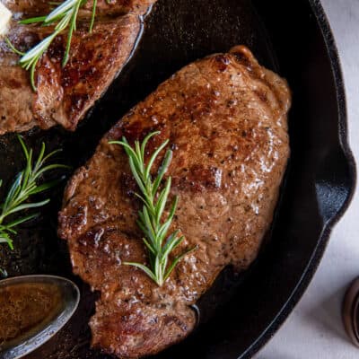 A top down shot of two steaks topped with sprigs of rosemary in a cast iron skillet.