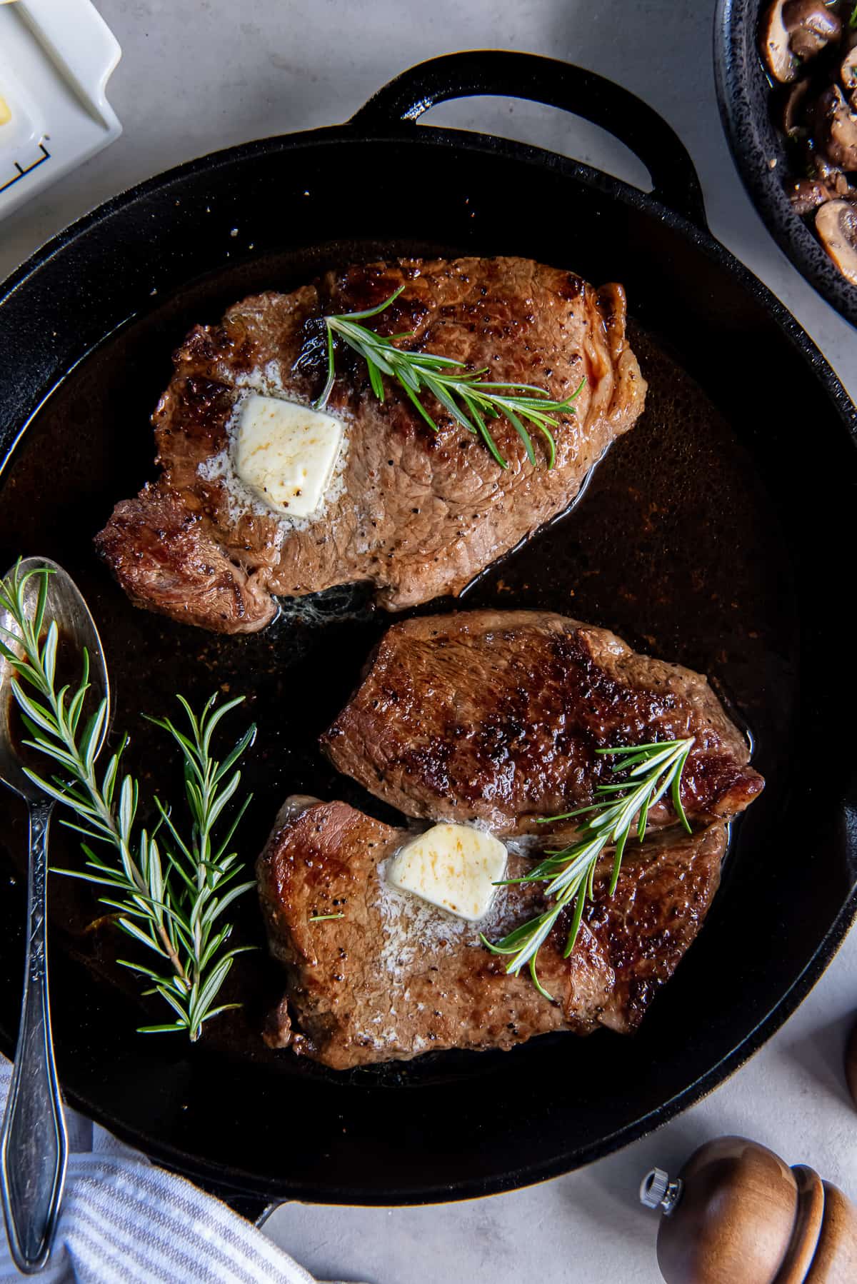 A top down shot of two steaks topped with a pat of butter and sprigs of rosemary in a cast iron skillet.