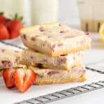 Three Strawberry Cheesecake Bars stacked on a wire rack with sliced strawberries next to them.