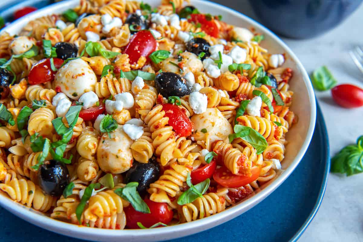 A large bowl filled with pasta salad with tomatoes, black olives, goat cheese, and basil.