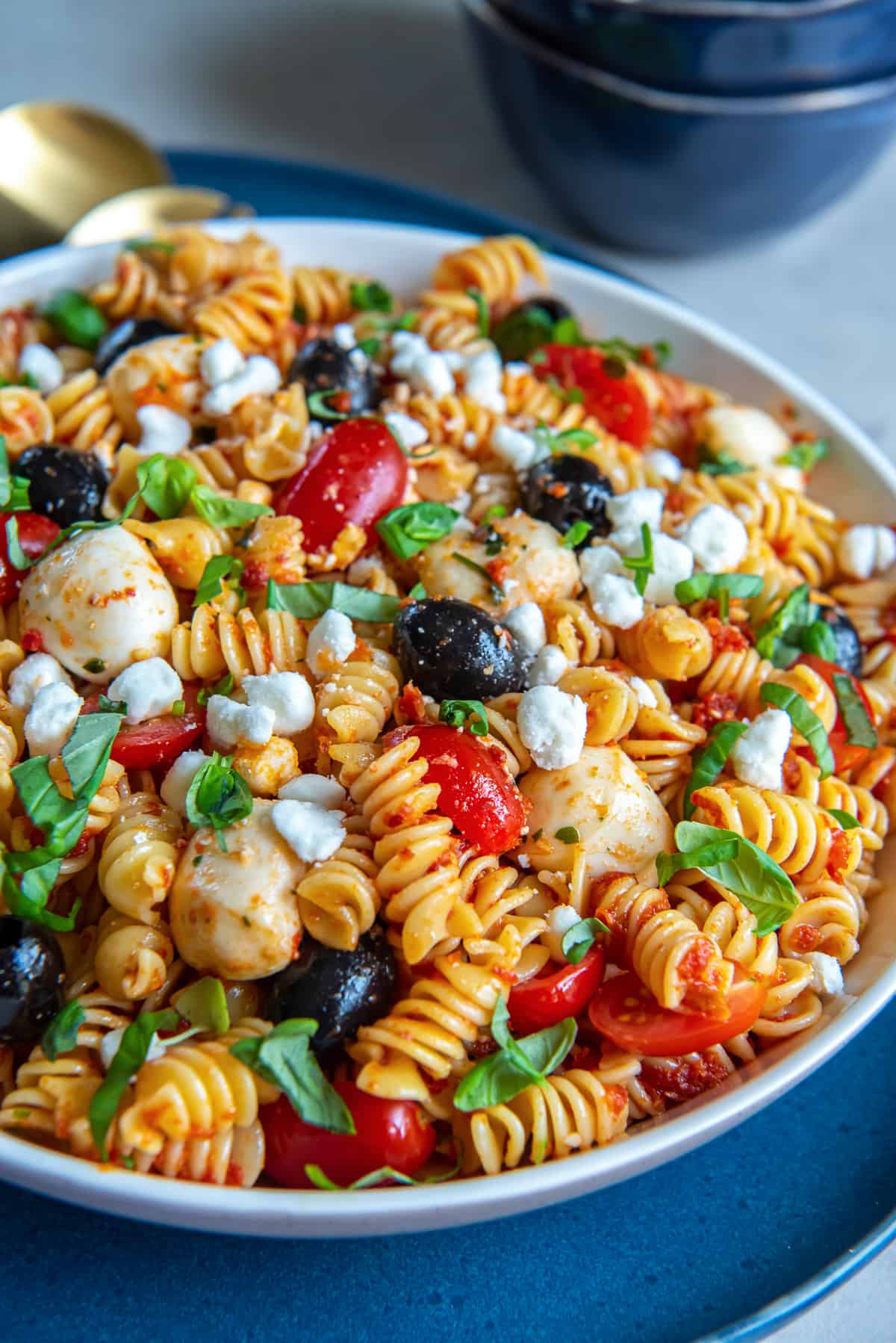 A side view of a large bowl filled with pasta salad with tomatoes, black olives, mozzarella balls, and basil.