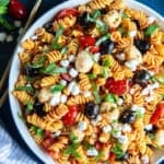 A top down shot of a large bowl on a blue platter filled with Sun-Dried Tomato Pasta Salad with gold salad spoons next to it.