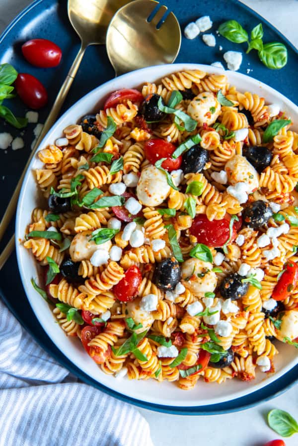 A top down shot of a large bowl on a blue platter filled with Sun-Dried Tomato Pasta Salad with gold salad spoons next to it.
