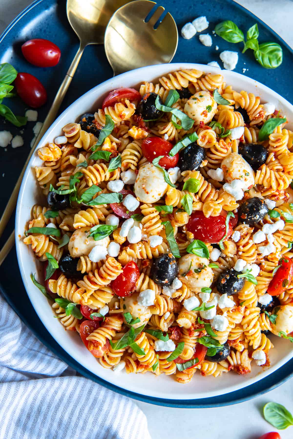 A top down shot of a large bowl on a blue platter filled with Sun-Dried Tomato Pasta Salad with gold salad spoons next to it.