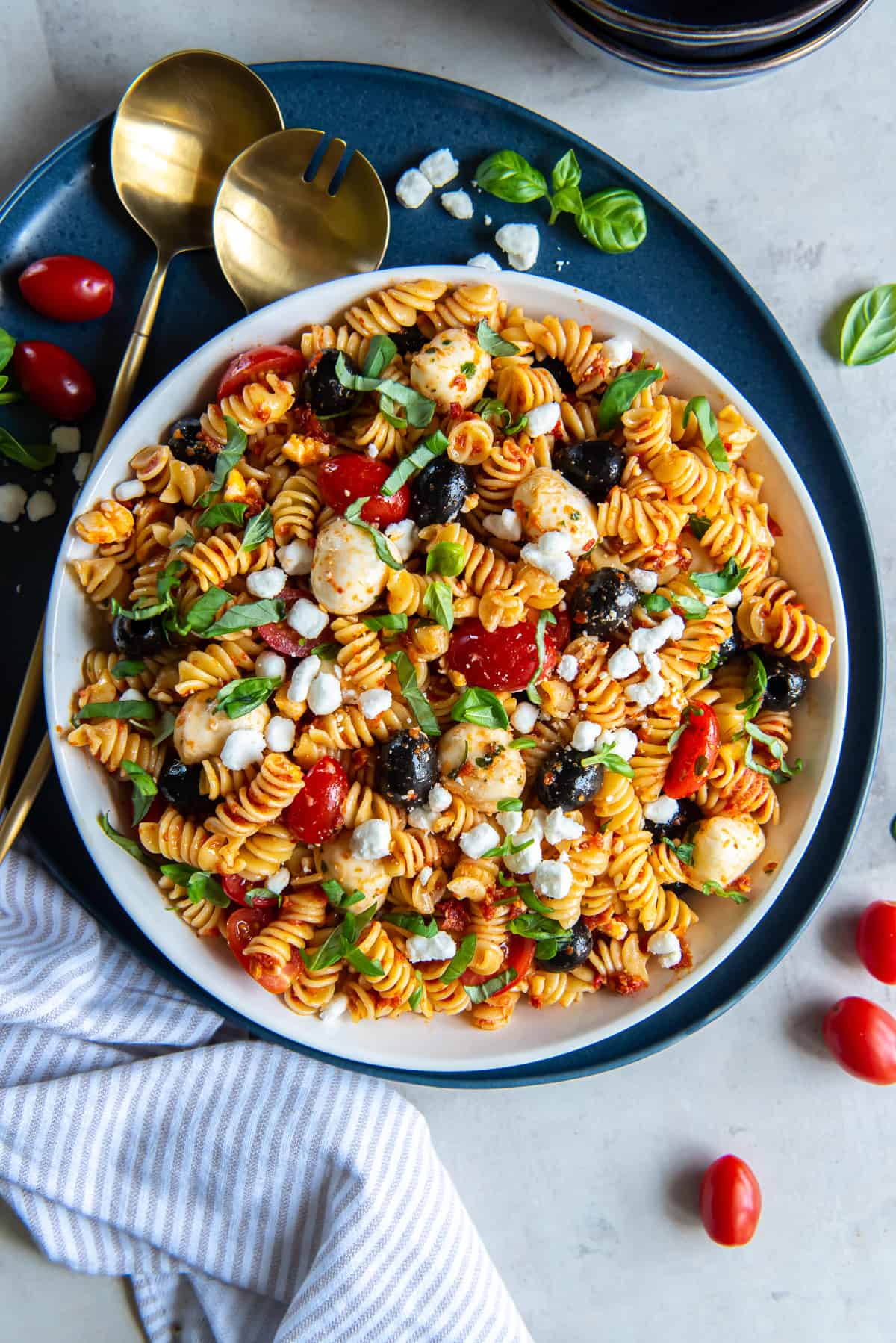 A top down shot of a large bowl on a blue platter filled with Sun-Dried Tomato Pasta Salad with gold salad spoons next to it.