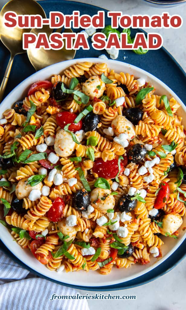 A top down shot of a large bowl on a blue platter filled with Sun-Dried Tomato Pasta Salad with gold salad spoons next to it with text.