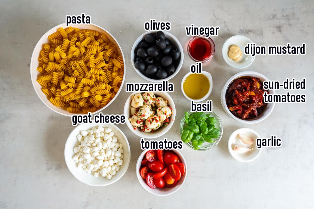 Dry fusilli pasta, olives, sun-dried tomatoes and other ingredients in bowls with text.