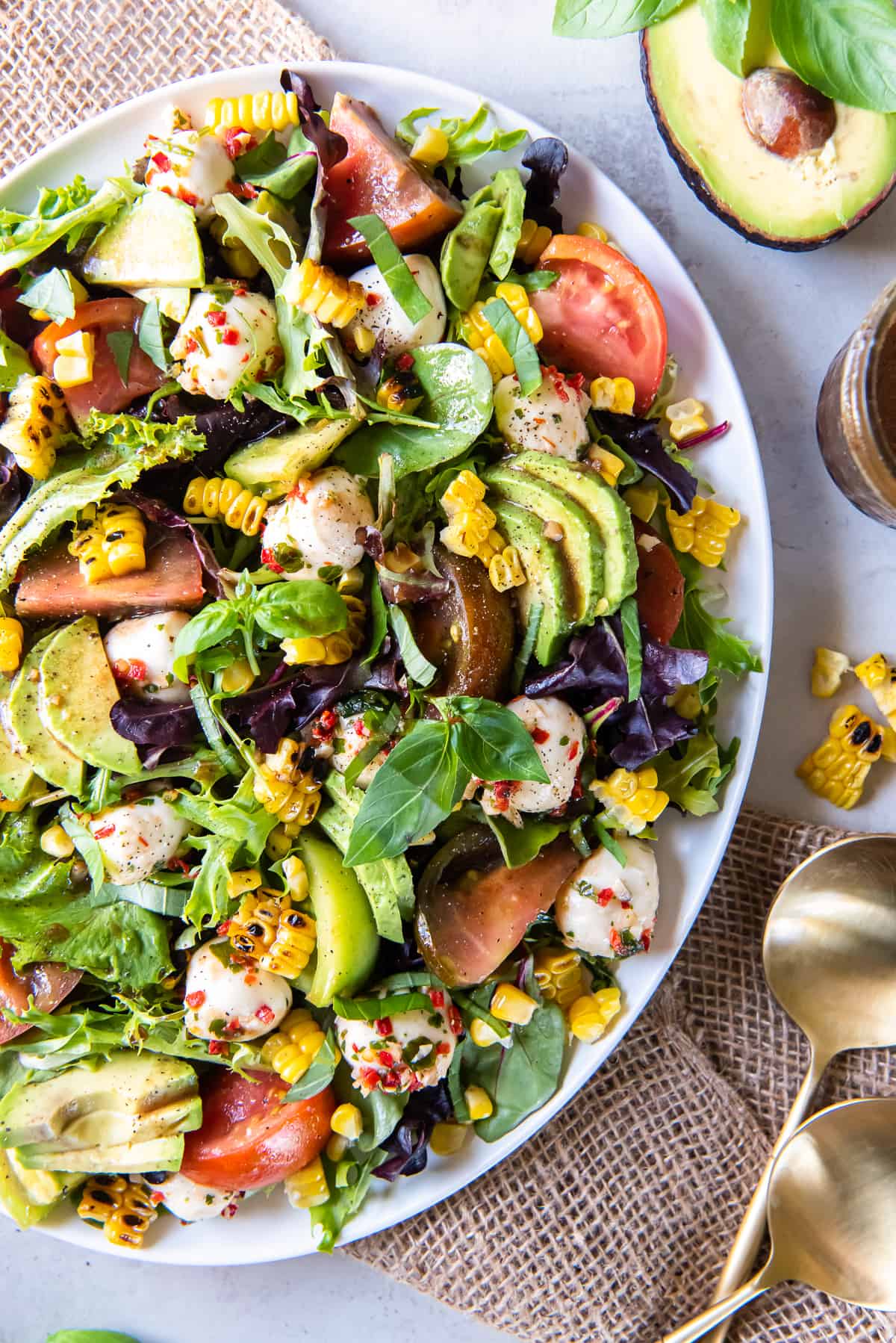 A top down shot of a caprese salad on top of a bed of greens with corn and avocado on a white platter surrounded by half an avocado and salad spoons.