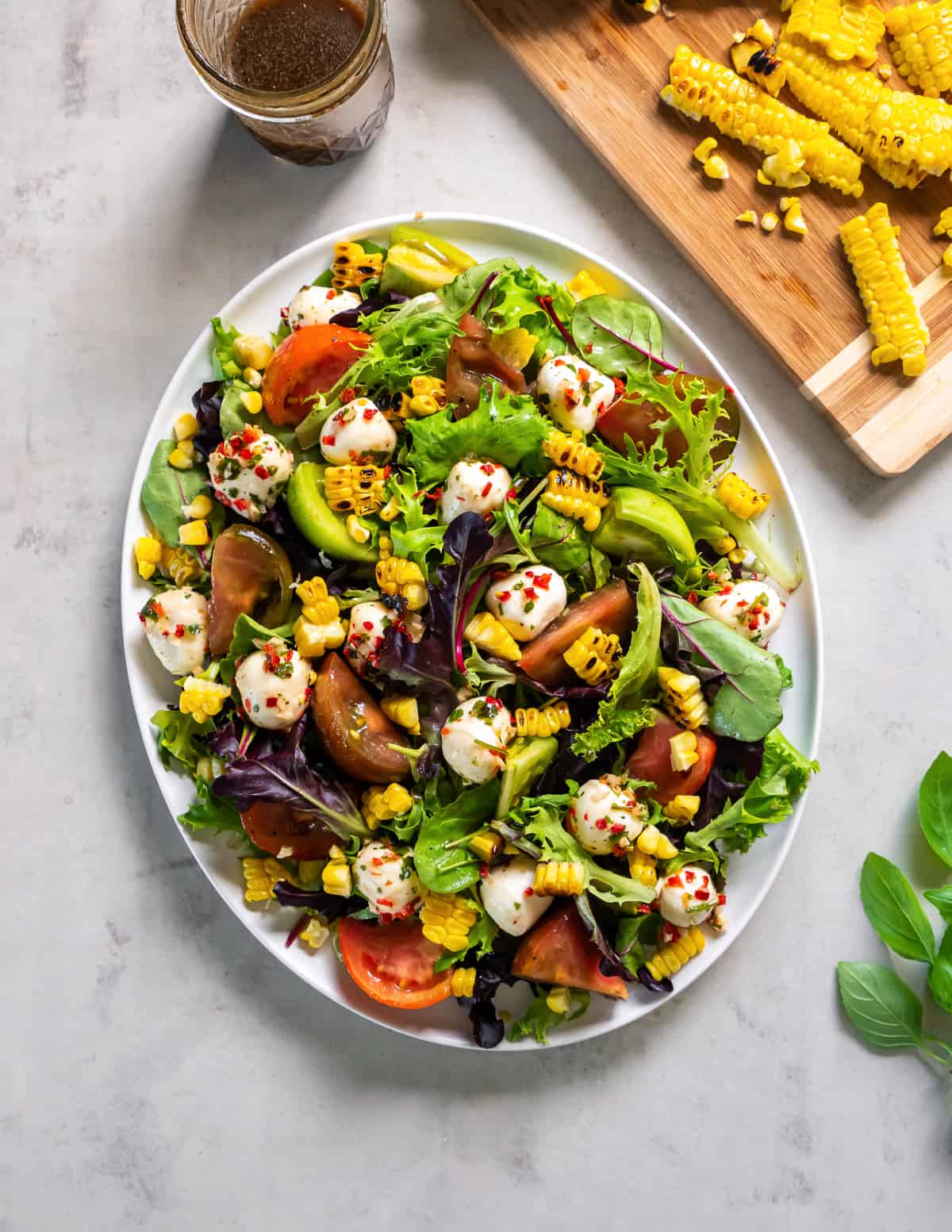 Caprese salad on a bed of greens with grilled corn and avocado layered on a white platter.