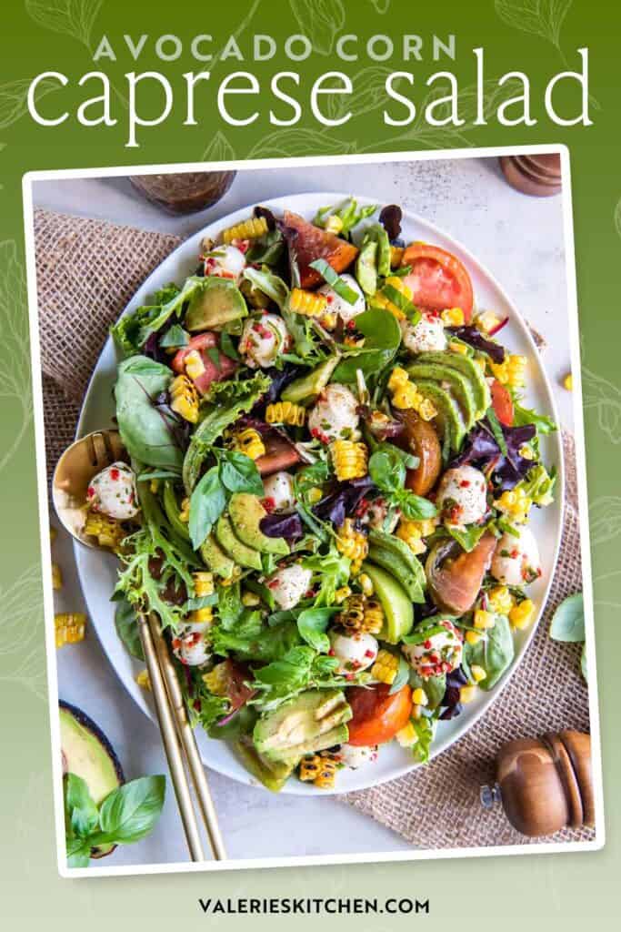 Serving spoons resting on a platter of avocado corn caprese salad with text overlay.