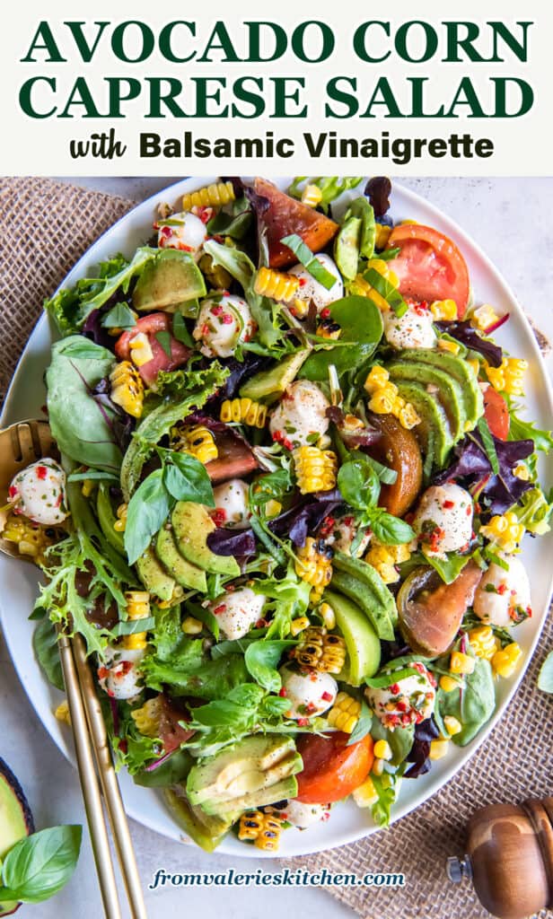 A top down shot of a caprese salad on top of a bed of greens with corn and avocado on a white platter with text.