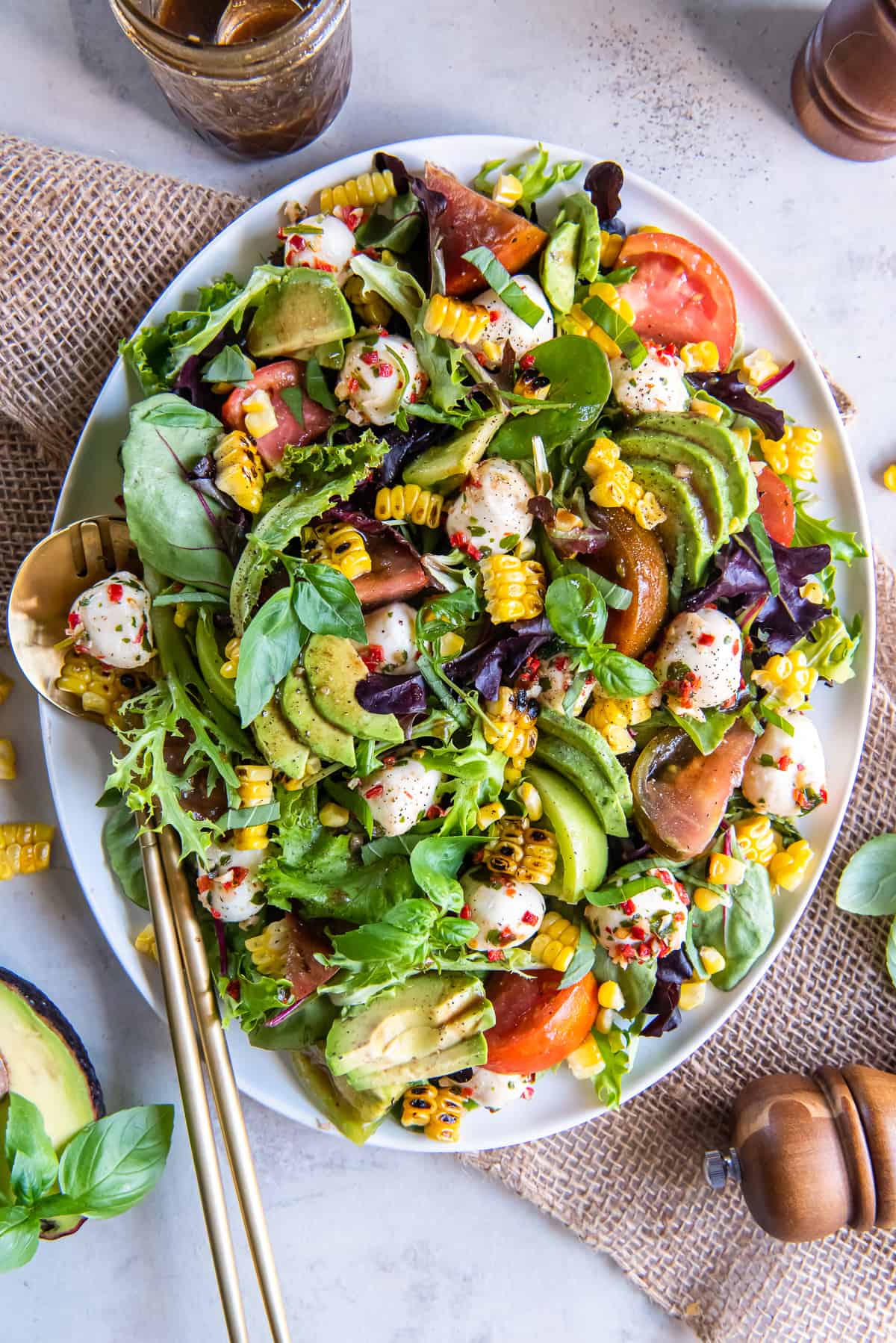 A top down shot of a caprese salad on top of a bed of greens with corn and avocado on a white platter.