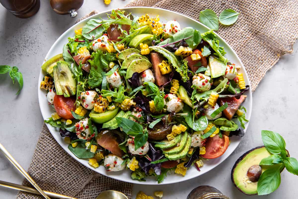 A top down shot of a caprese salad on top of a bed of greens with corn and avocado on a white platter surrounded by half an avocado and basil leaves.