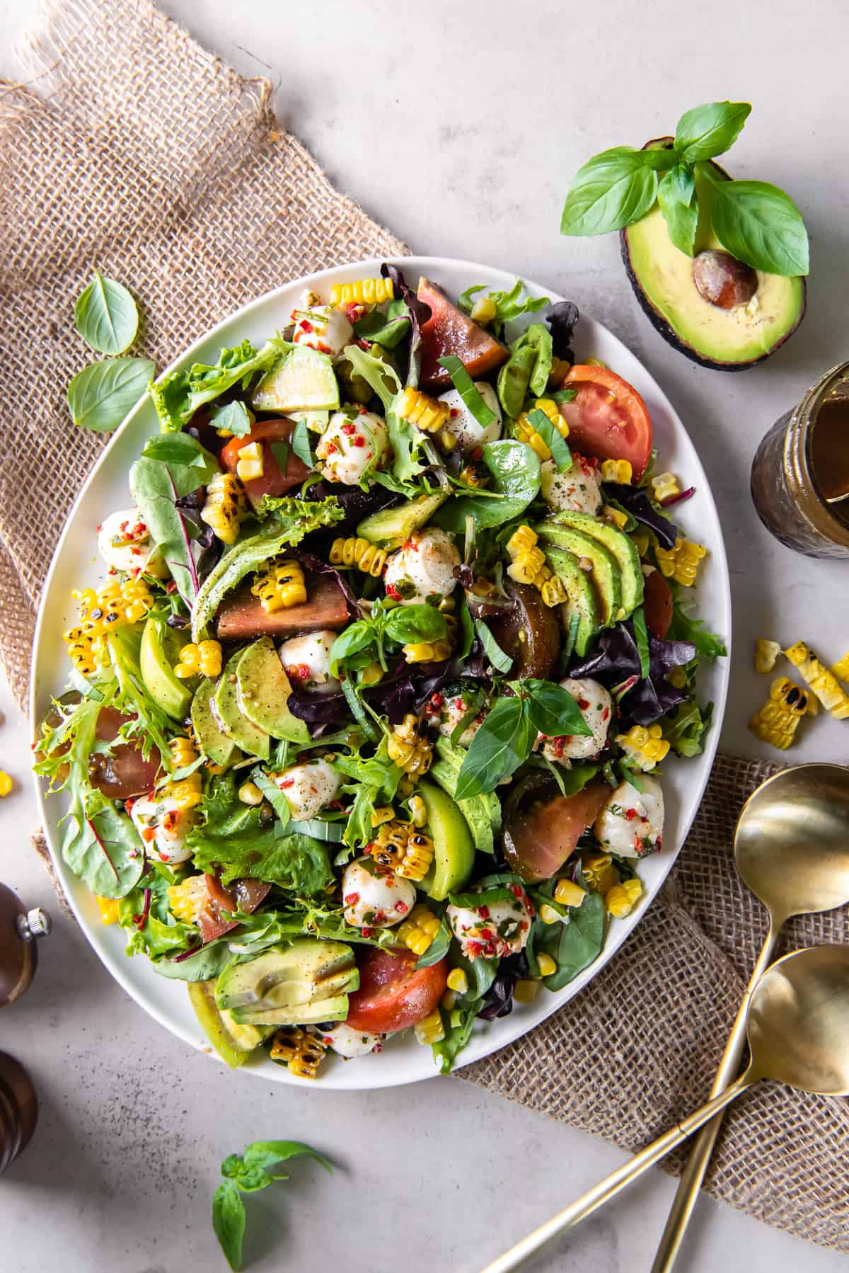 A top down shot of a caprese salad on top of a bed of greens with corn and avocado on a white platter.