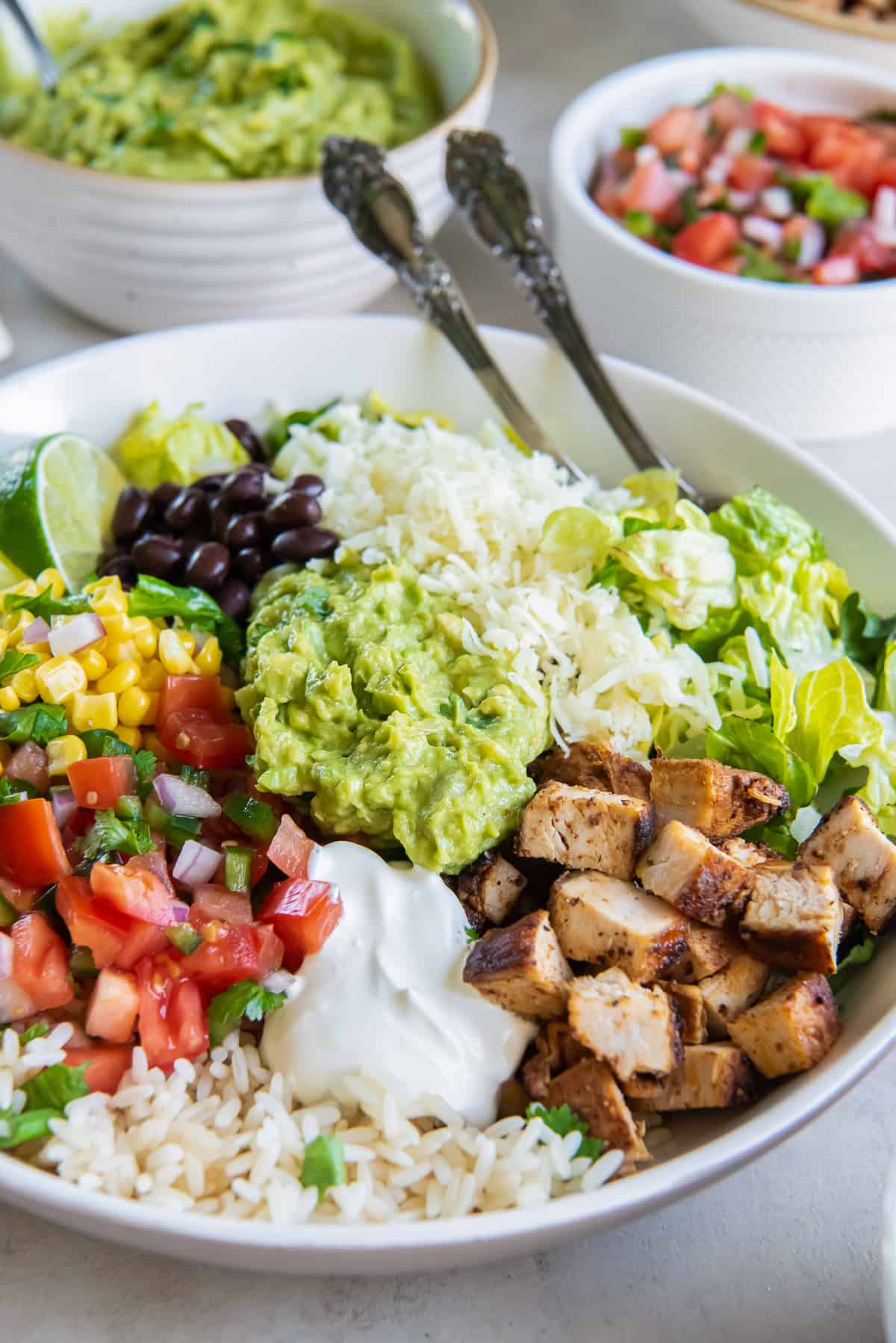 A chicken burrito bowl in a white bowl with forks and small bowls of salsa and guacamole behind it.