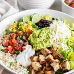 A chicken burrito bowl in a white bowl and small bowls of salsa and guacamole behind it.
