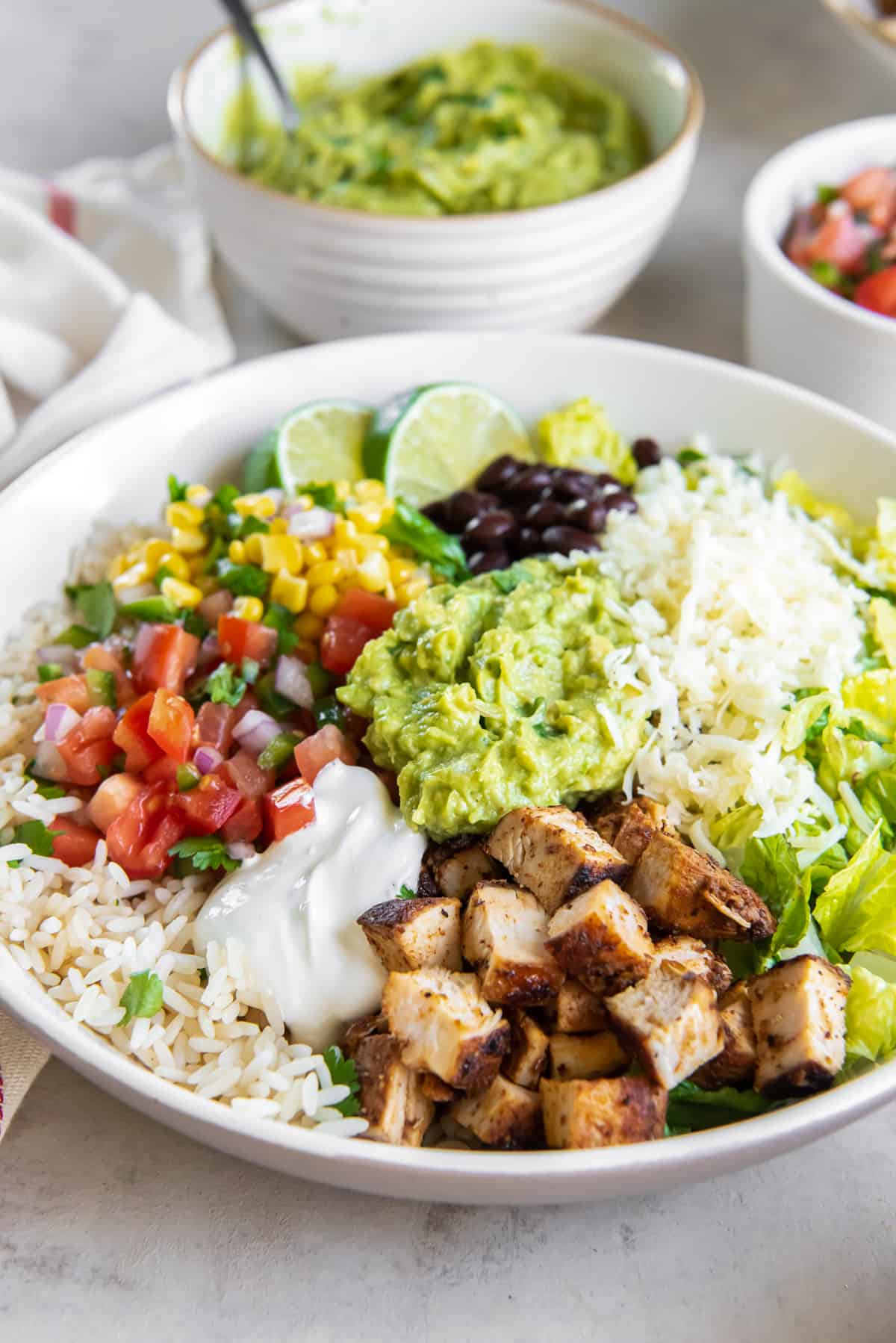 A chicken burrito bowl in a white bowl and small bowls of salsa and guacamole behind it.