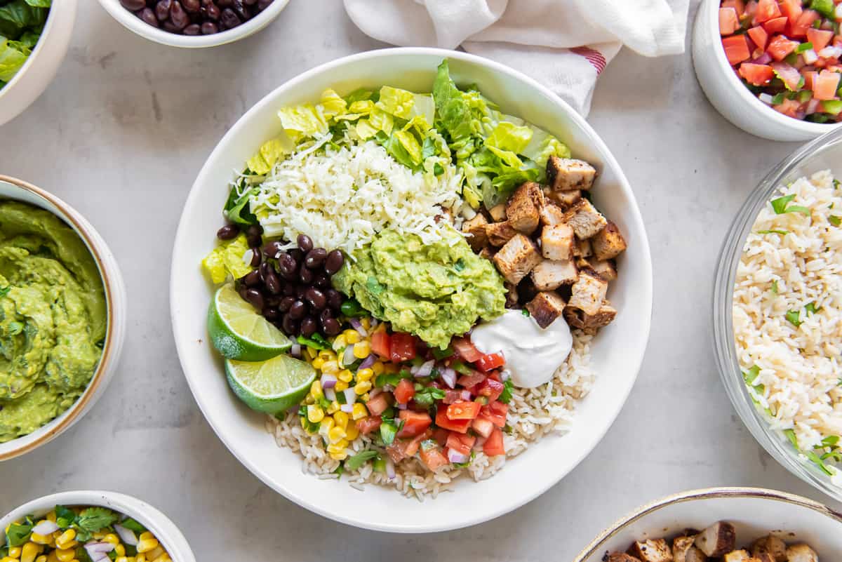 A top down shot of a copycat Chipotle burrito bowl with chicken next to small bowls filled with ingredients.