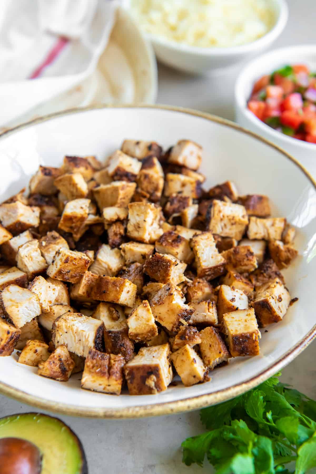 A side view of cubed copycat Chipotle chicken in a white bowl surrounded by ingredients in smaller bowls.