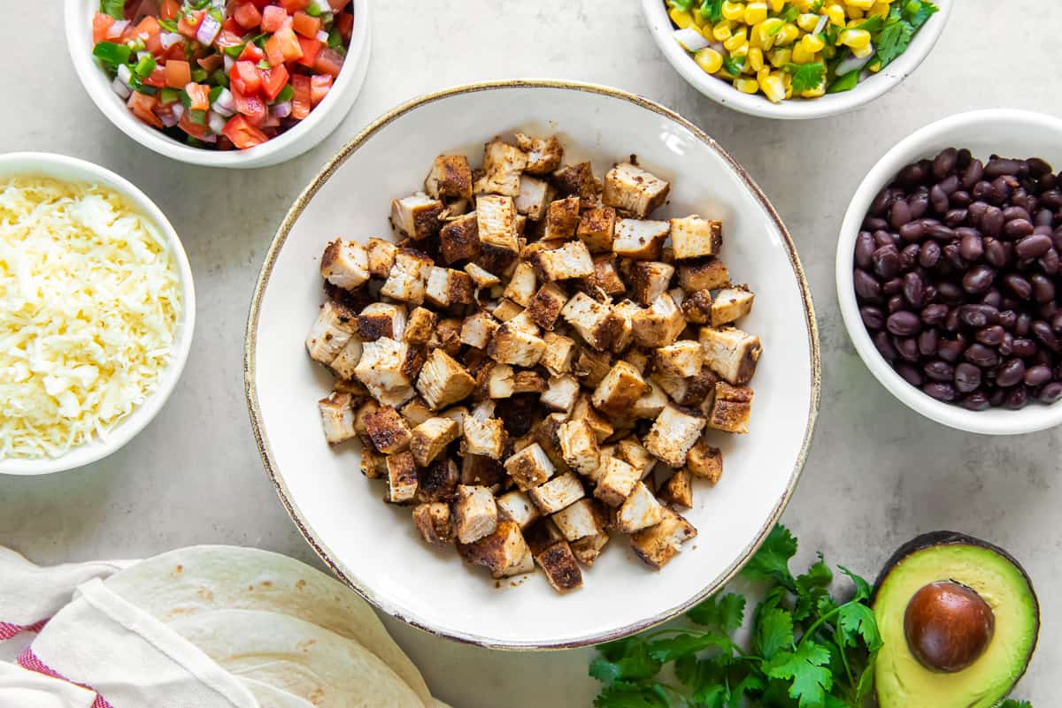 A bowl of copycat Chipotle Chicken surrounded by bowls filled with black beans, salsa, and other ingredients.