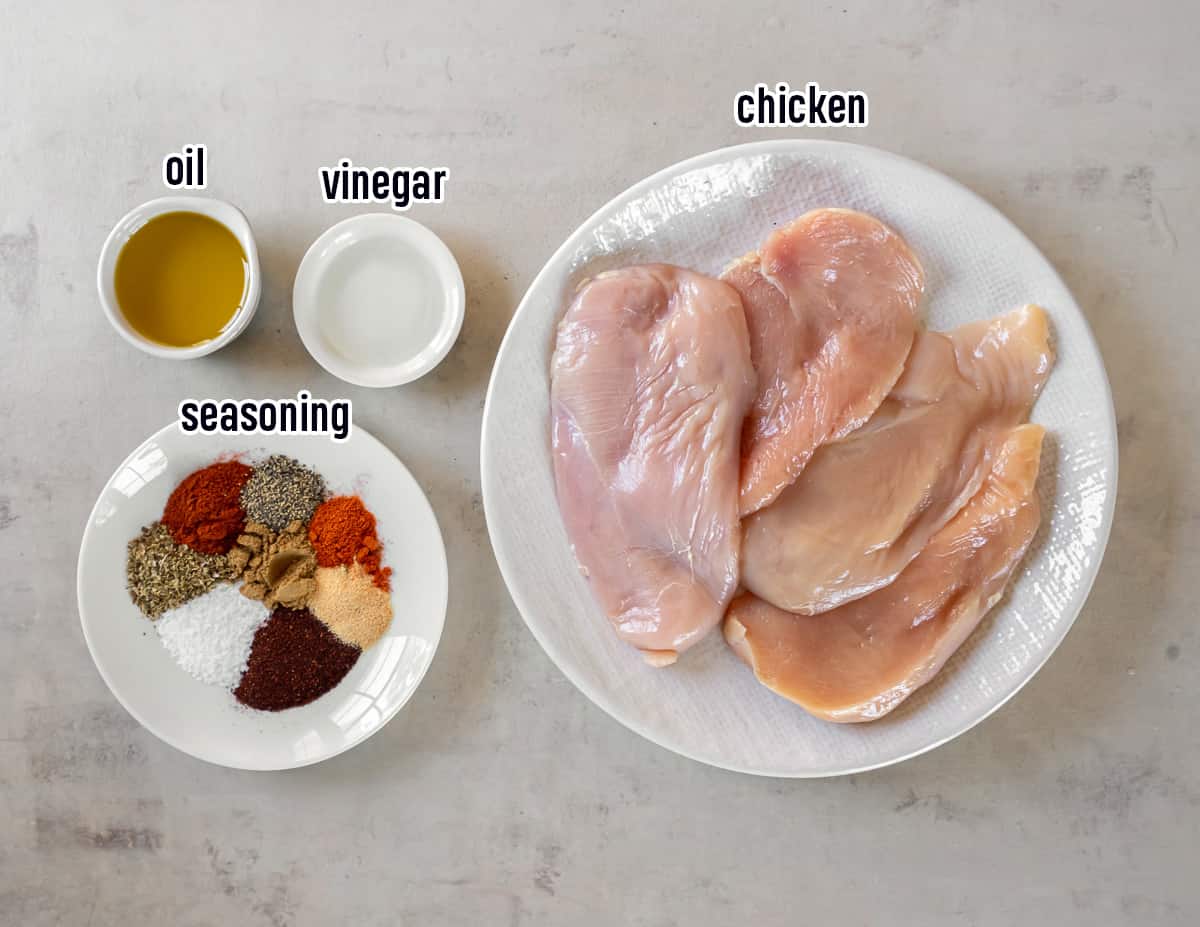 Boneless chicken breasts on a white plate next spices, oil, and vinegar in small bowls with text.