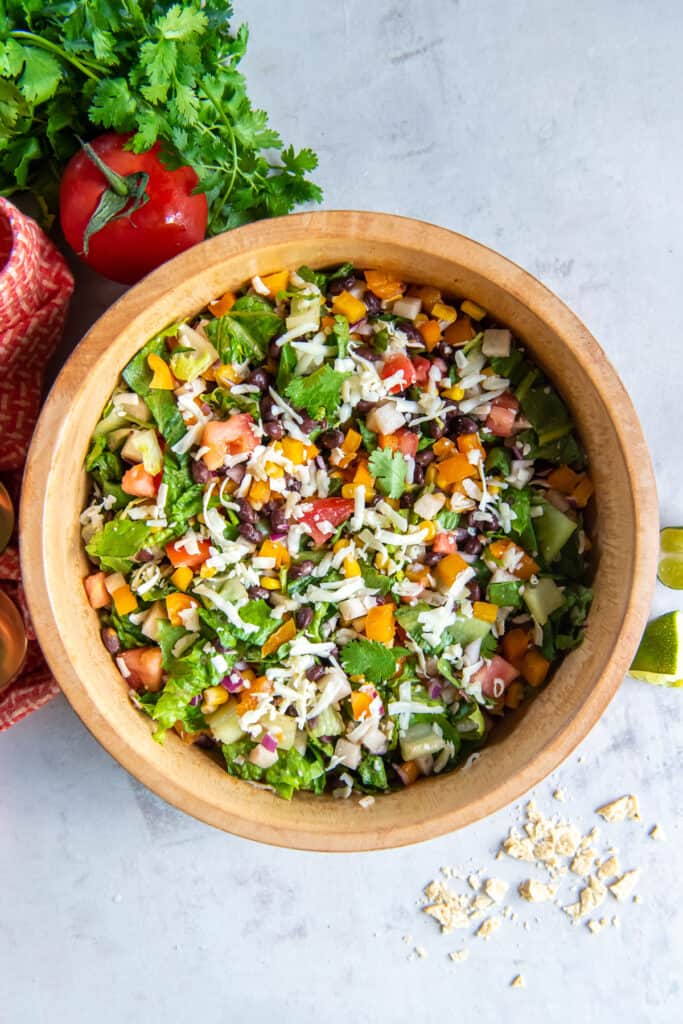 A top down shot of a chopped salad in a wood bowl next to a tomato and sprigs of cilantro.