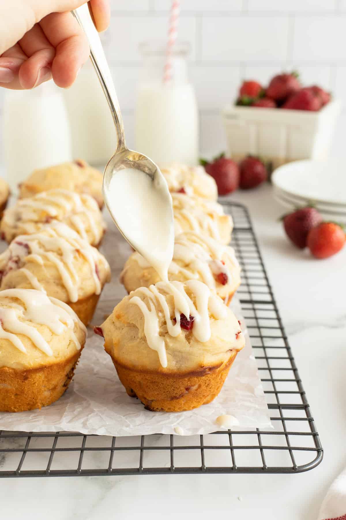 A spoon drizzling icing over a strawberry muffin on a wire rack.