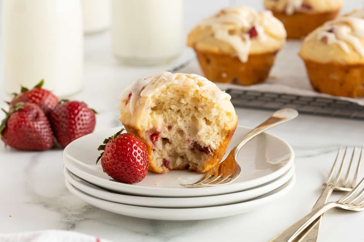 A strawberry muffin with a bite missing on a stack of white plates with a fresh strawberry and a fork.