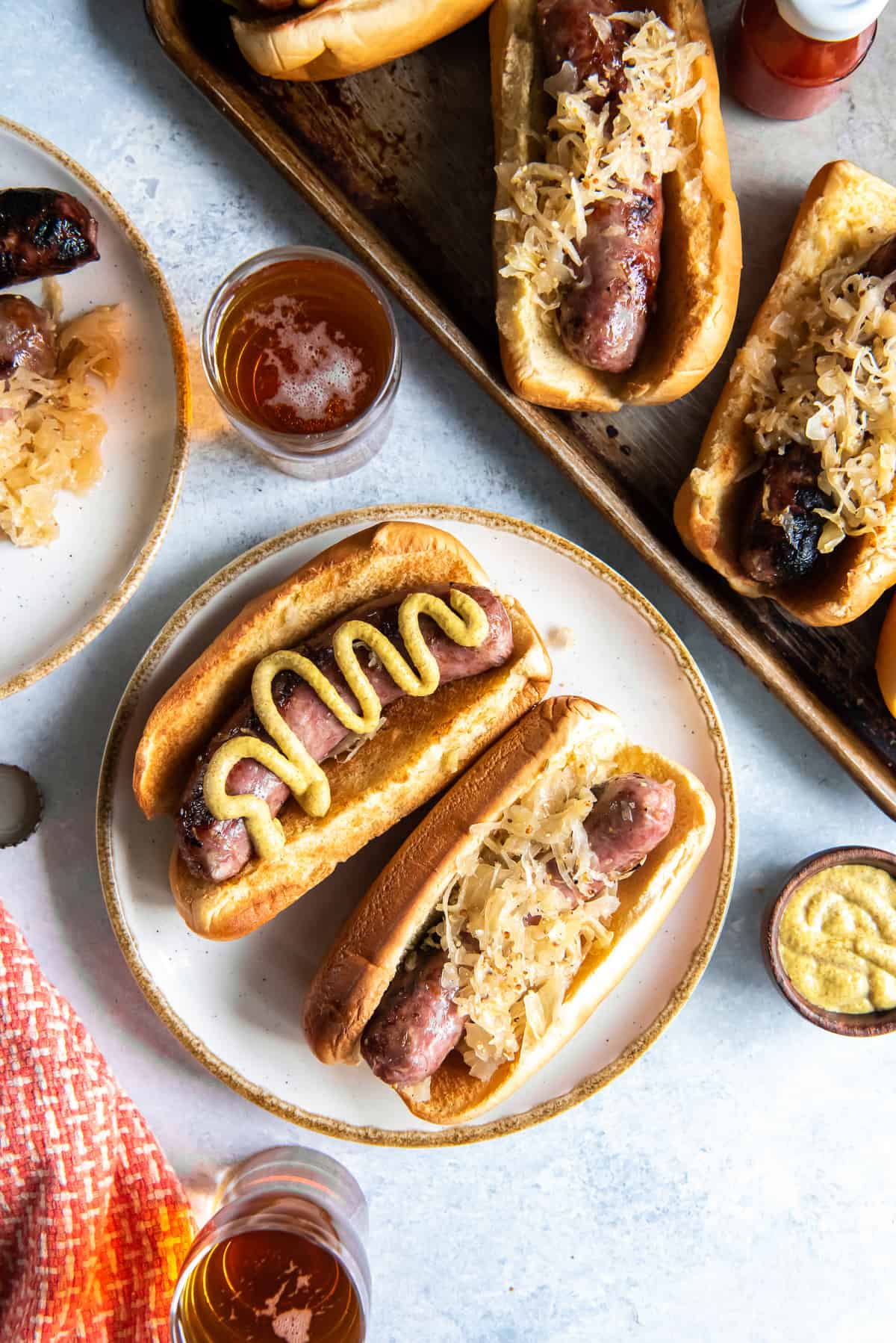 A top down shot of two beer brats on buns with sauerkraut and mustard on a white plate next to a baking sheet full.